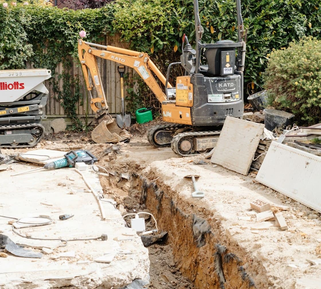 Mini excavator digging a trench beside a house in a fenced backyard